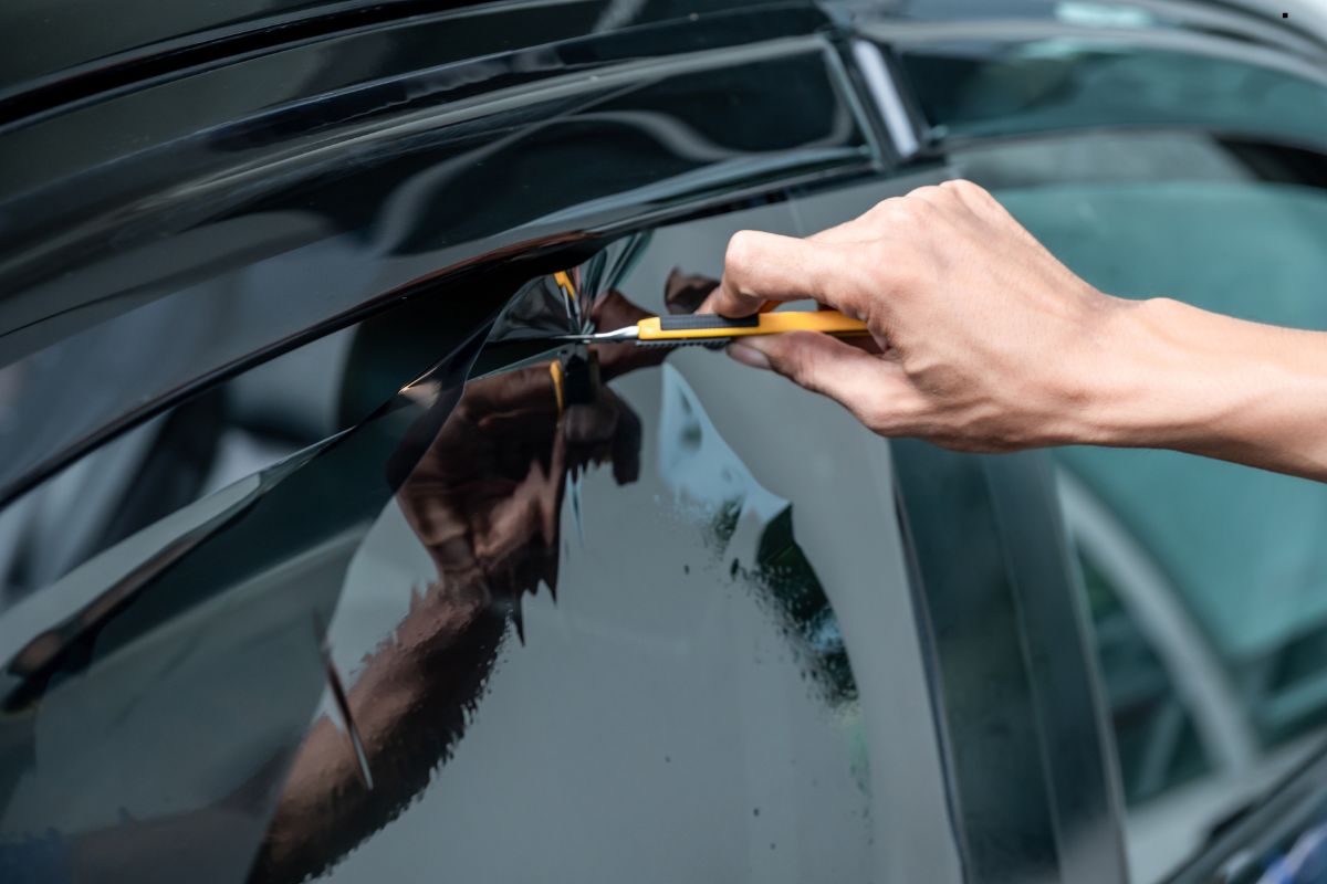 A person uses a utility knife to trim black vinyl wrap on a car window, focusing on the precise cut along the edge