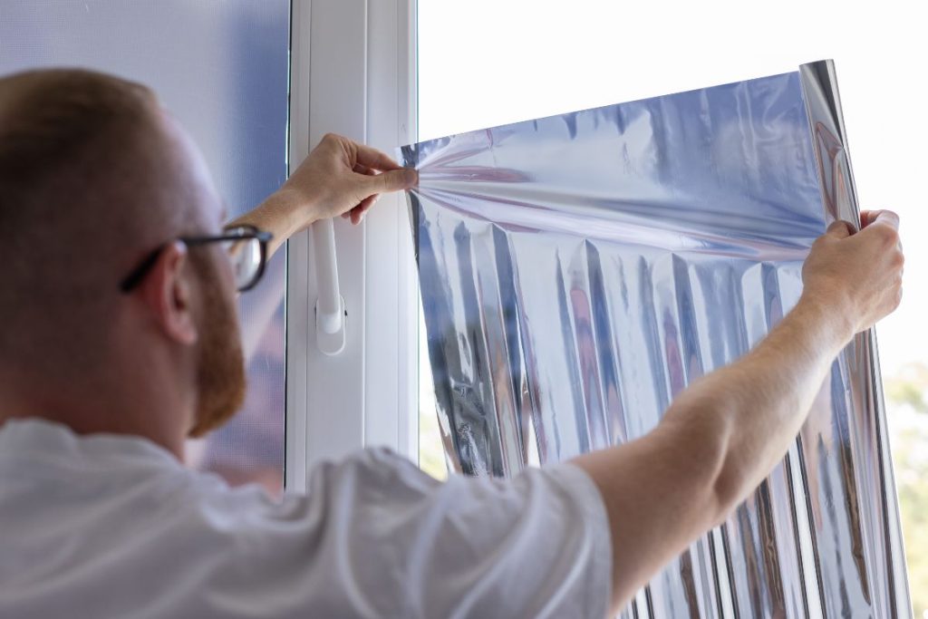 A man applies aluminum foil to a window, likely for insulation or to block sunlight.