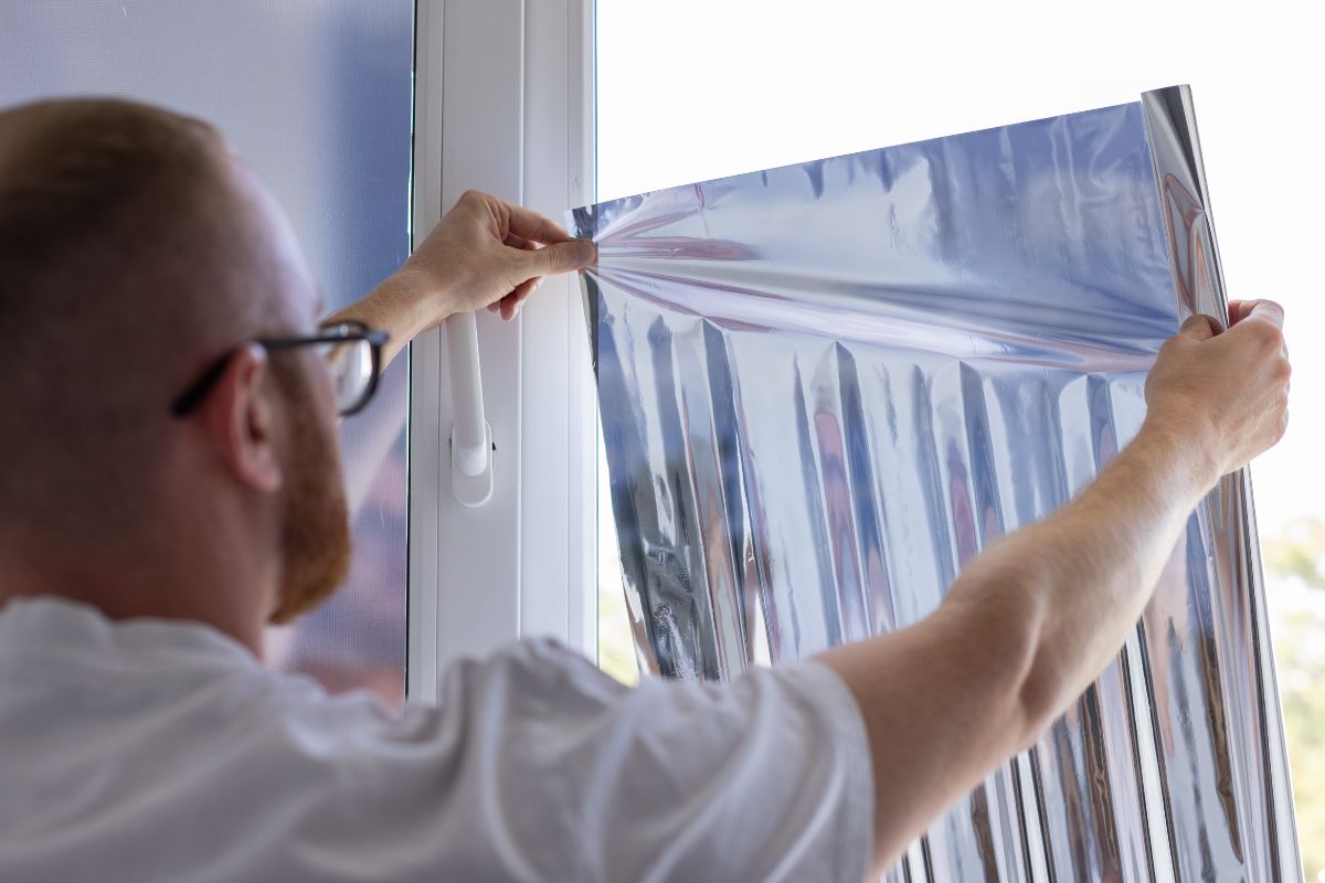 A man applies aluminum foil to a window, likely for insulation or to block sunlight.